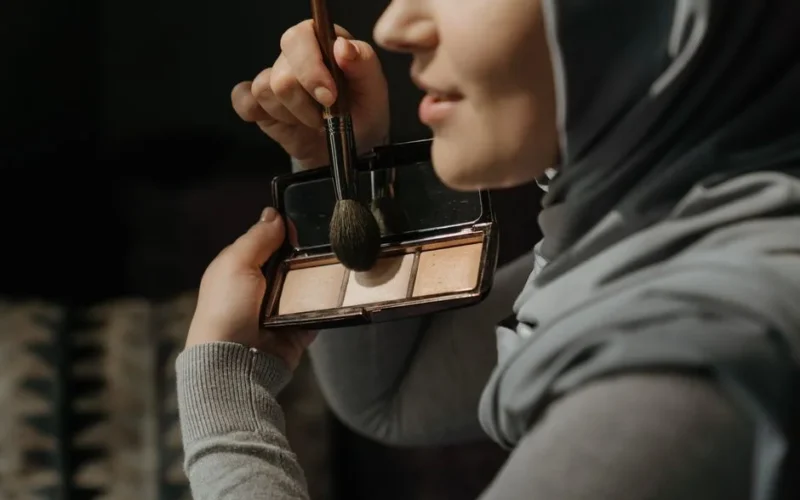 A close-up of a woman in a hijab applying makeup with a brush indoors.