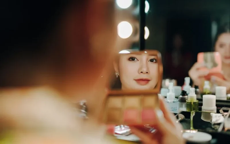 A woman applying makeup, captured in a mirror reflection surrounded by beauty products.