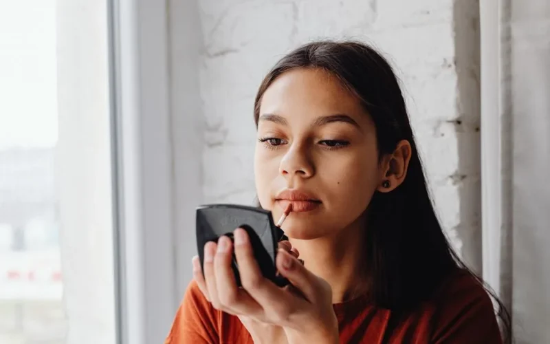 A young woman applying lip tint while looking into a mirror indoors by a window.