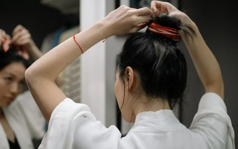 Woman in bathrobe styling hair in front of mirror, focusing on hair bun.
