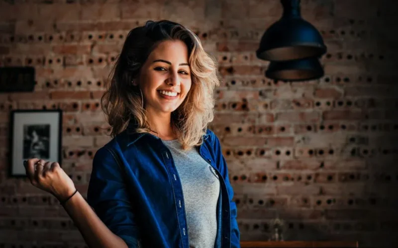 Portrait of a smiling woman in denim jacket against a brick wall indoors.