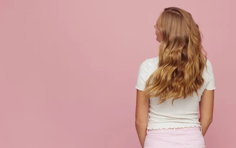 Rear view of woman with long blonde wavy hair against pink backdrop.