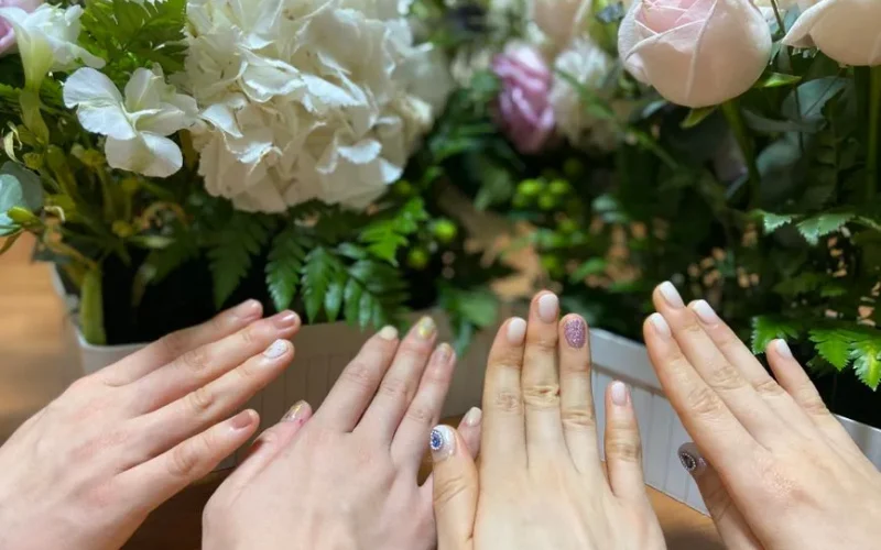 Group of hands with varied nail art surrounded by lush flower arrangements in pastel tones.