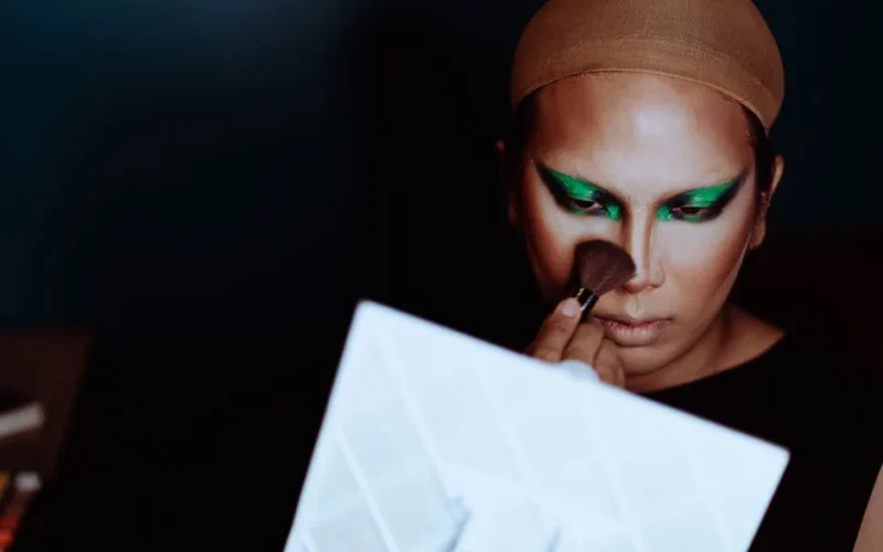 Crop young ethnic female theater artist with bright eye shadow on eyelids using cosmetic brush in front of mirror