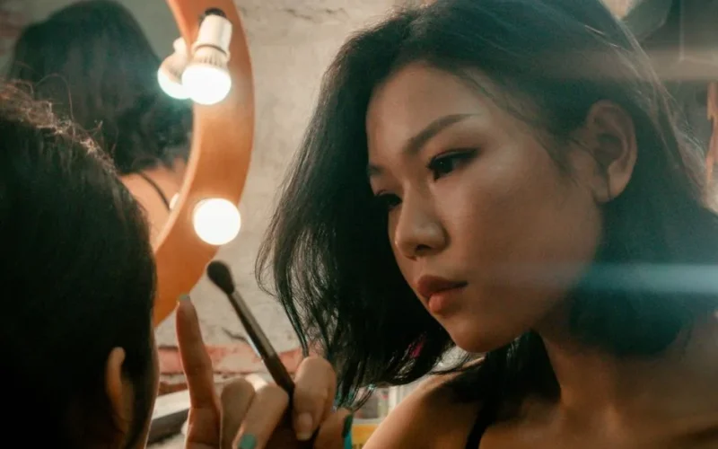 Asian woman using a makeup brush, applying makeup in front of a mirror.