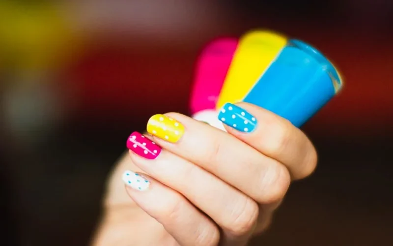 Close-up of a hand with colorful polka dot nail art holding vibrant nail polish bottles.