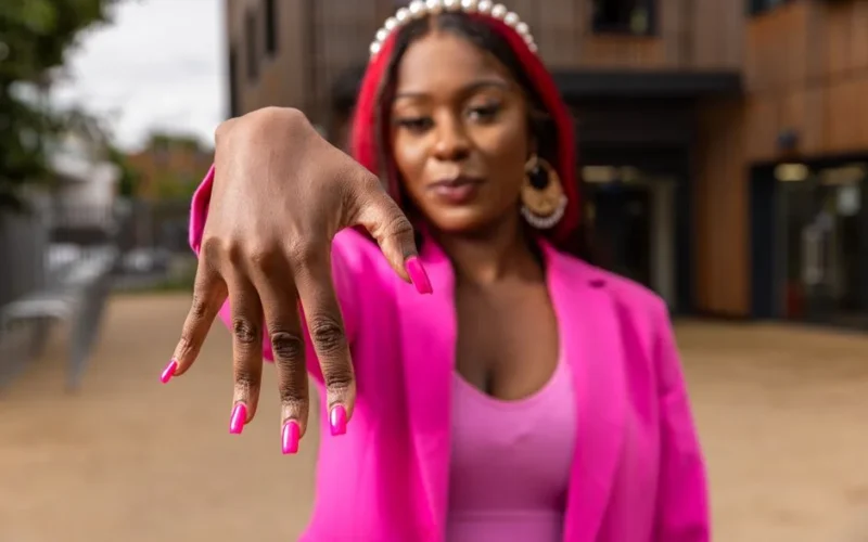 Confident woman with pink nails and outfit displaying stylish manicure outdoors.