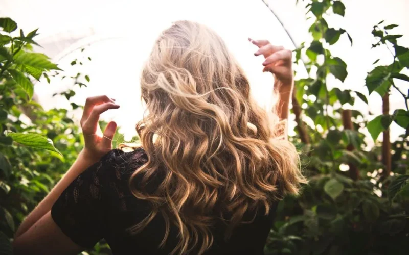 Blonde woman with curly hair surrounded by lush greenery, enjoying a sunny day.