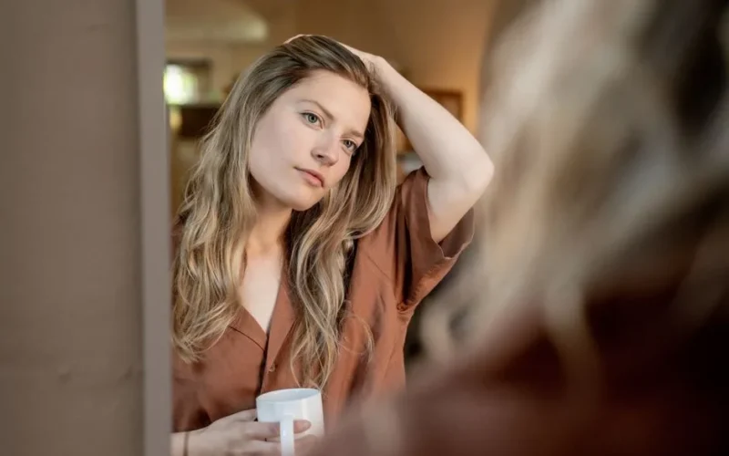 A woman gazes into a mirror, sipping coffee at home, with a serene expression and natural light accentuating her features.