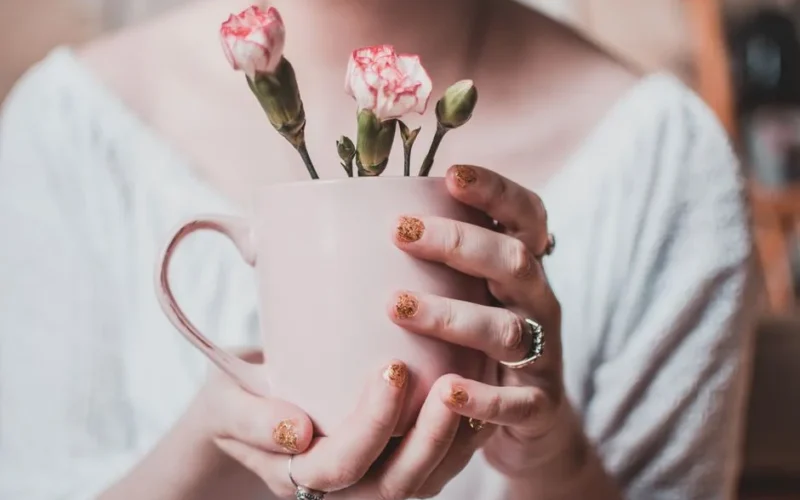 Aesthetic pink carnation buds in a ceramic mug, gently held by a woman with glittery nails.