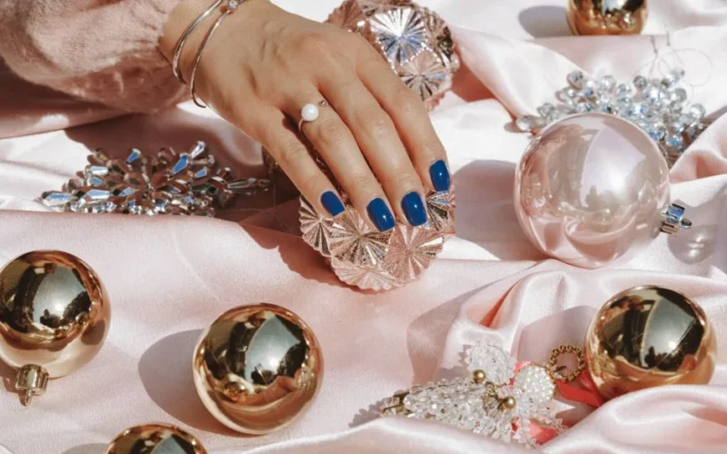 Stylish hand showcasing blue nails resting on pink satin surrounded by opulent ornaments and jewelry.