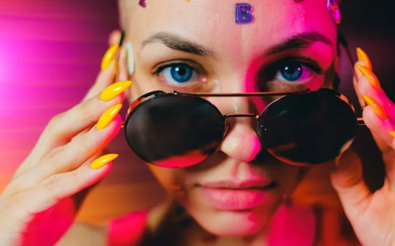 Crop bald female with multicolored stickers and bright manicure lowering stylish sunglasses and looking at camera in studio