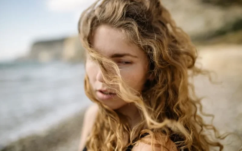 Young woman with curly blonde hair on a windy beach day.