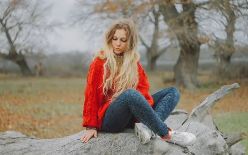 A young woman with long blonde hair sits on a log outdoors, wearing a red sweater, jeans, and sneakers.