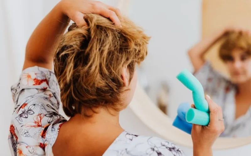 Woman using curlers to style hair in front of a bathroom mirror, focusing on wellness and self-care.