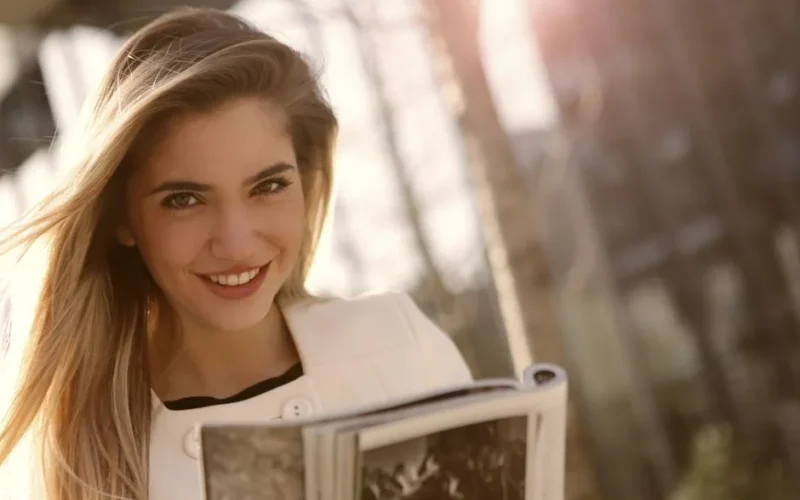 Smiling woman with long blond hair reading a magazine outdoors in sunlight.