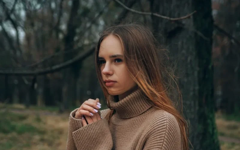 A contemplative woman poses in a woodland setting, wearing a cozy brown sweater on a fall day.