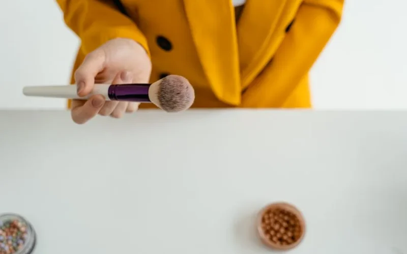 A hand holding a makeup brush over a table with cosmetics, featuring a yellow jacket.