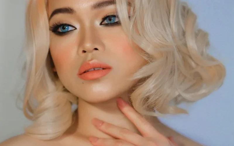 Blonde woman with striking blue eyes and elegant makeup posing in a studio portrait.