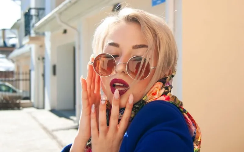Trendy blond woman with sunglasses and floral scarf posing outdoors, showcasing chic fashion and manicured nails.