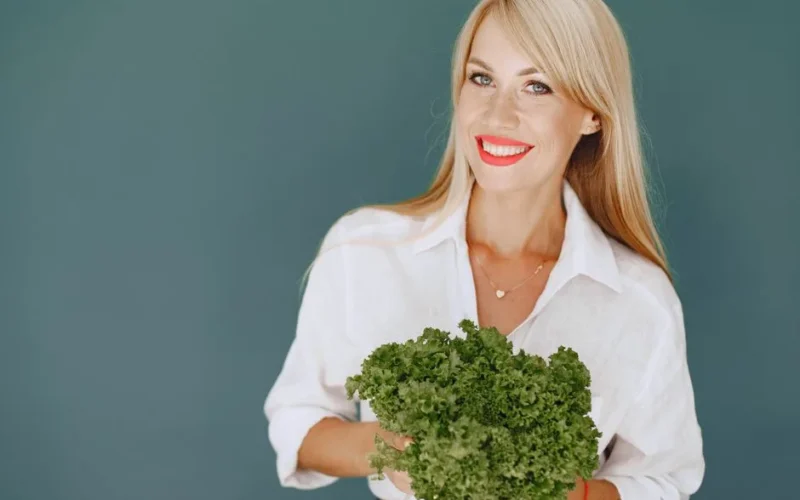 Cheerful woman with blonde hair holding fresh lettuce, promoting healthy living in a studio setting.