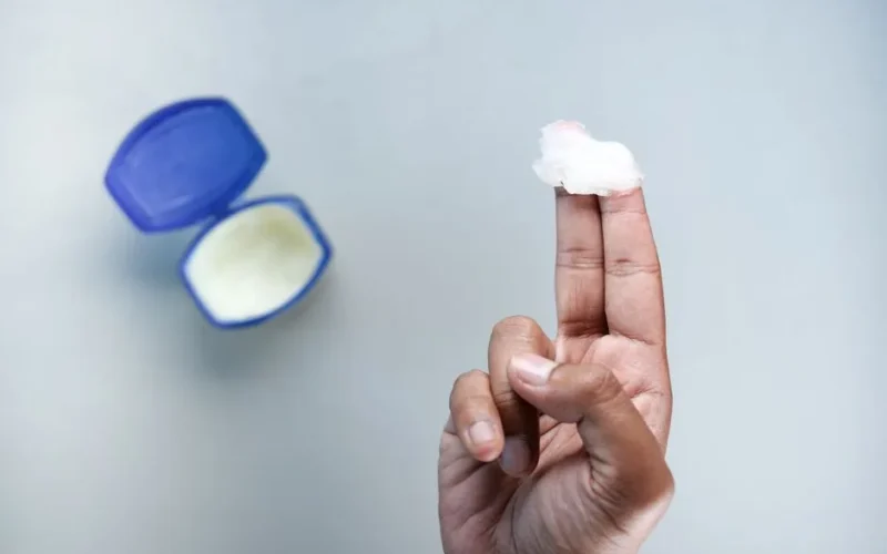 A hand holding petroleum jelly on fingers, next to an open container. Skin care concept.