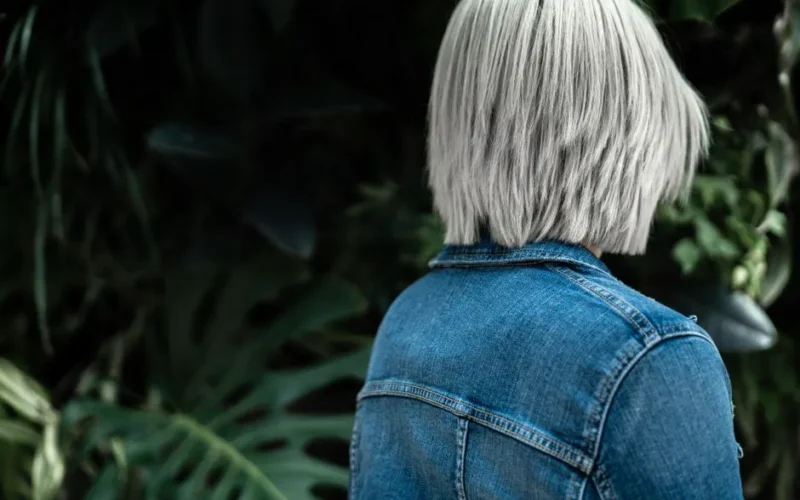 Woman with white hair in denim jacket surrounded by lush greenery outdoors.
